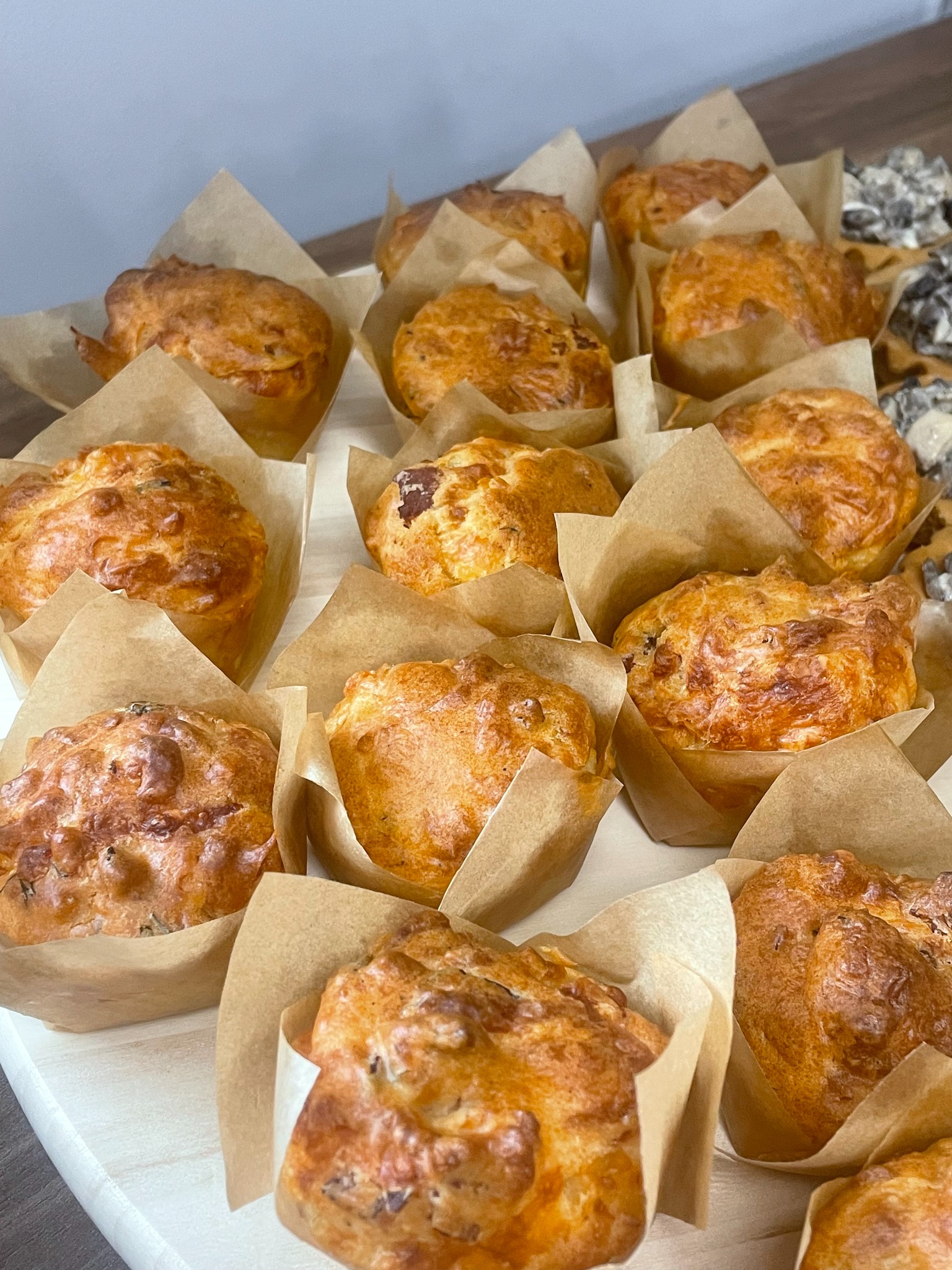 Apetyczne muffiny wytrawne w papierowych papilotkach, ułożone rzędami na białej desce. Widoczne kawałki mięsa i ziół. Idealne na catering firmowy lub świąteczny.