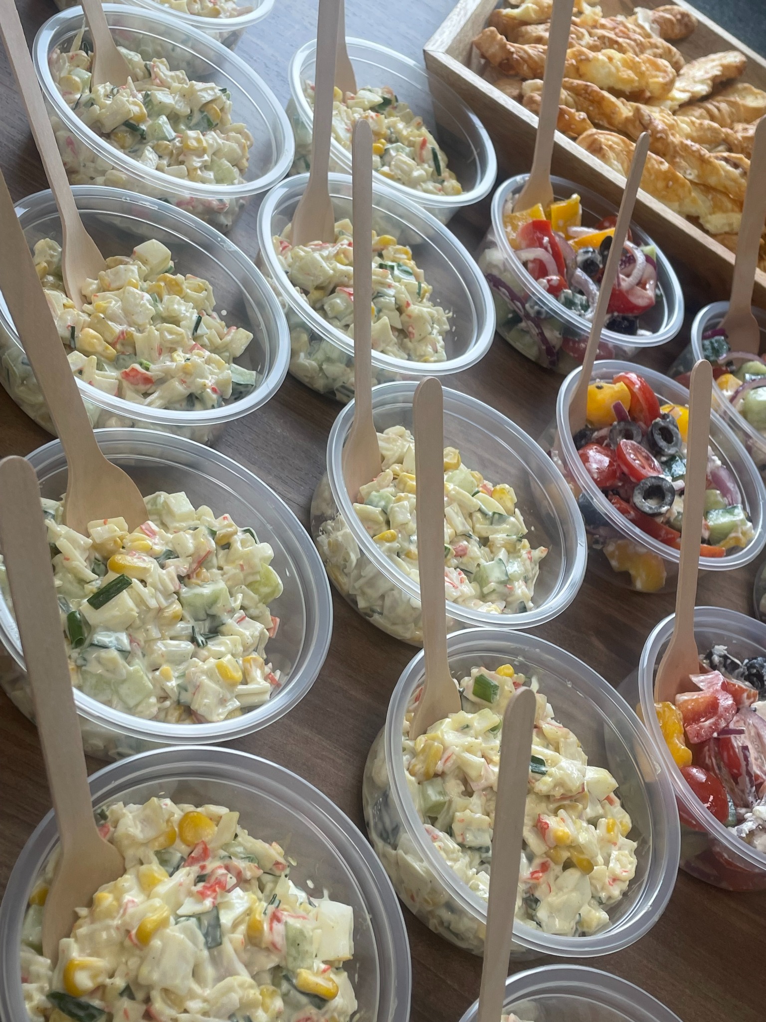 Różnorodne sałatki w plastikowych pojemnikach z drewnianymi łyżkami, ułożone na drewnianym stole. W tle przekąski w drewnianej tacy. Przygotowane porcje.