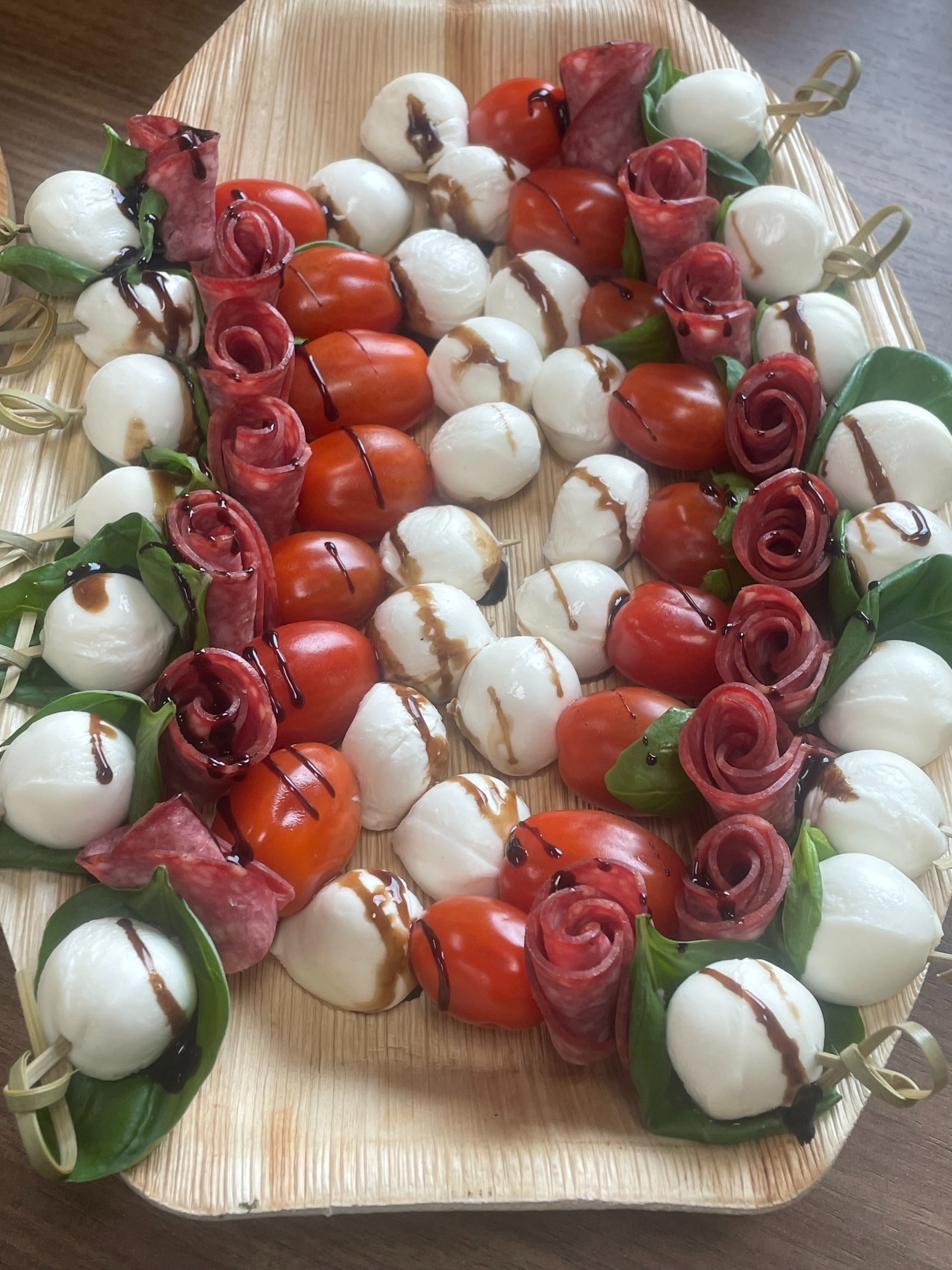 Elegancka kompozycja przekąsek: mini-szaszłyki z pomidorków, mozzarelli, salami w kształcie różyczek, bazylii, polane kremem balsamicznym, ułożone na drewnianej tacy.