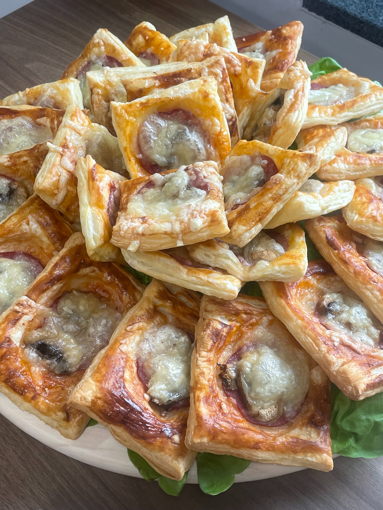 Kruche, kwadratowe przekąski z ciasta francuskiego z szynką, pieczarkami i serem, ułożone na tacy z zielonymi dekoracjami. Idealne na catering.