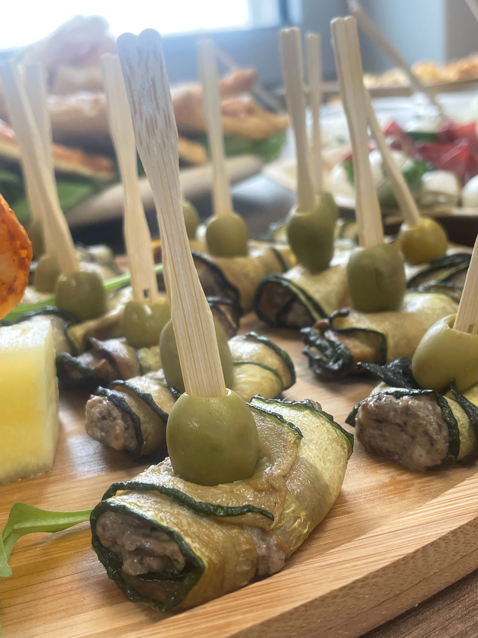 Roladki z cukinii z oliwką na drewnianej desce, przekąski cateringowe. Widoczne inne potrawy w tle, naturalne światło.