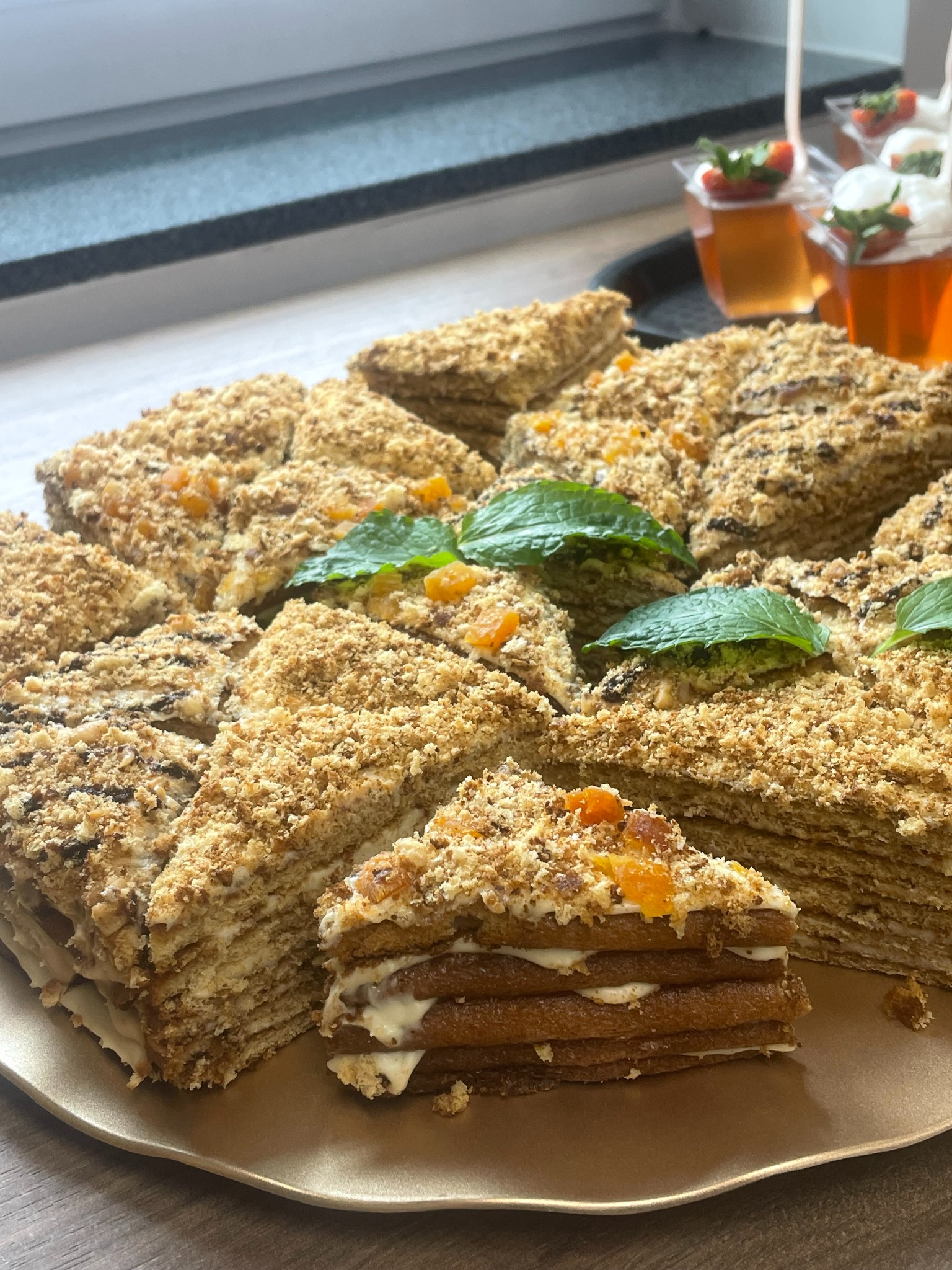 Krojony tort miodownik na złotej tacy, udekorowany miętą i morelami, z deserami w tle. Widoczne warstwy kremu i posypka z orzechów. Blat kuchenny w tle.