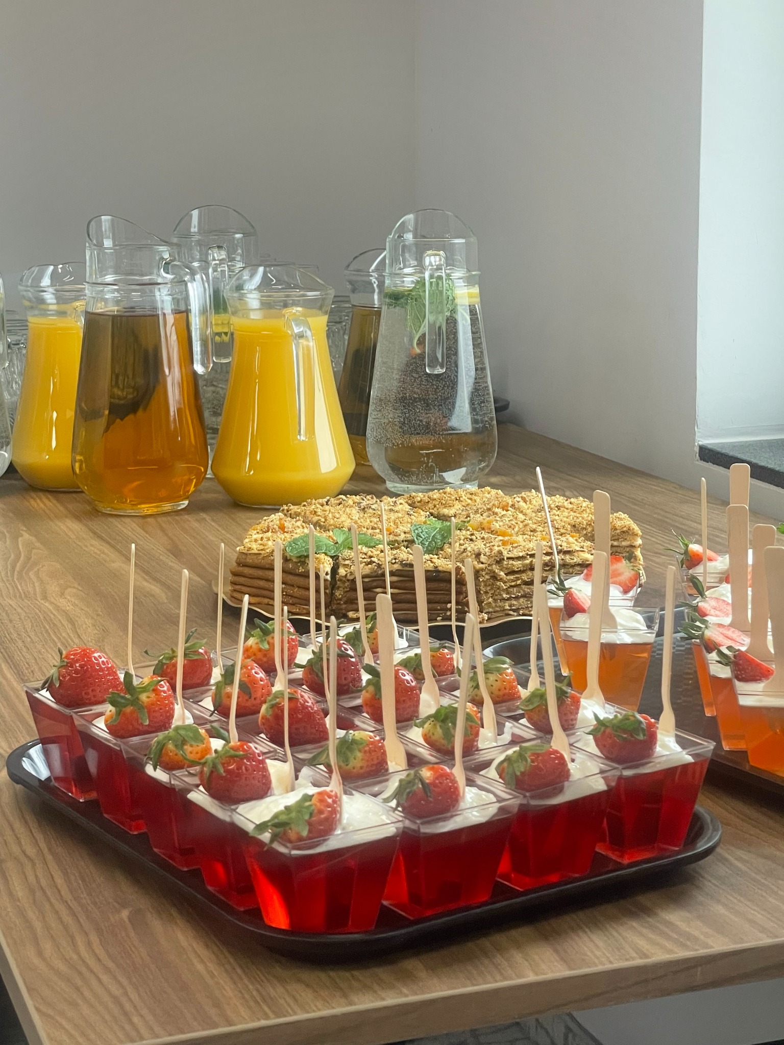 Stół cateringowy w Katowicach: dzbanki z napojami, tort orzechowy i galaretki z bitą śmietaną i truskawkami na tackach. Słodki bufet.