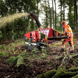 ArborTechLine - Operator w odzieży ochronnej podaje gałąź do czerwono-czarnego rębaka na gąsienicach, który wyrzuca zrębki w lesie. W tle wysokie drzewa.