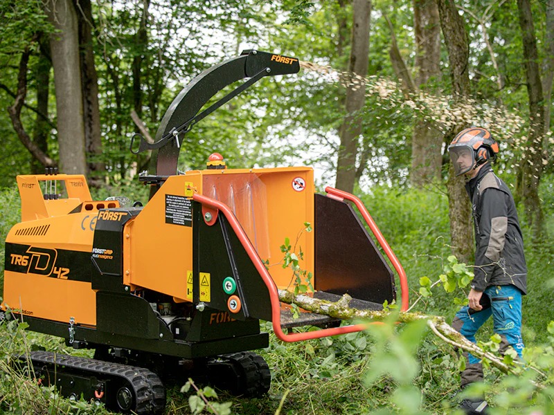 Pomarańczowa rębarka Forst TR6D42 w akcji, z operatorem w kasku i siatce ochronnej, rozdrabniająca gałęzie na tle zielonego lasu. Wióry w powietrzu.