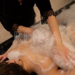 Reina - Kobieta relaksuje się podczas zabiegu z pianą, wykonywanego przez masażystkę w czarnym stroju. Delikatne oświetlenie podkreśla atmosferę odprężenia.