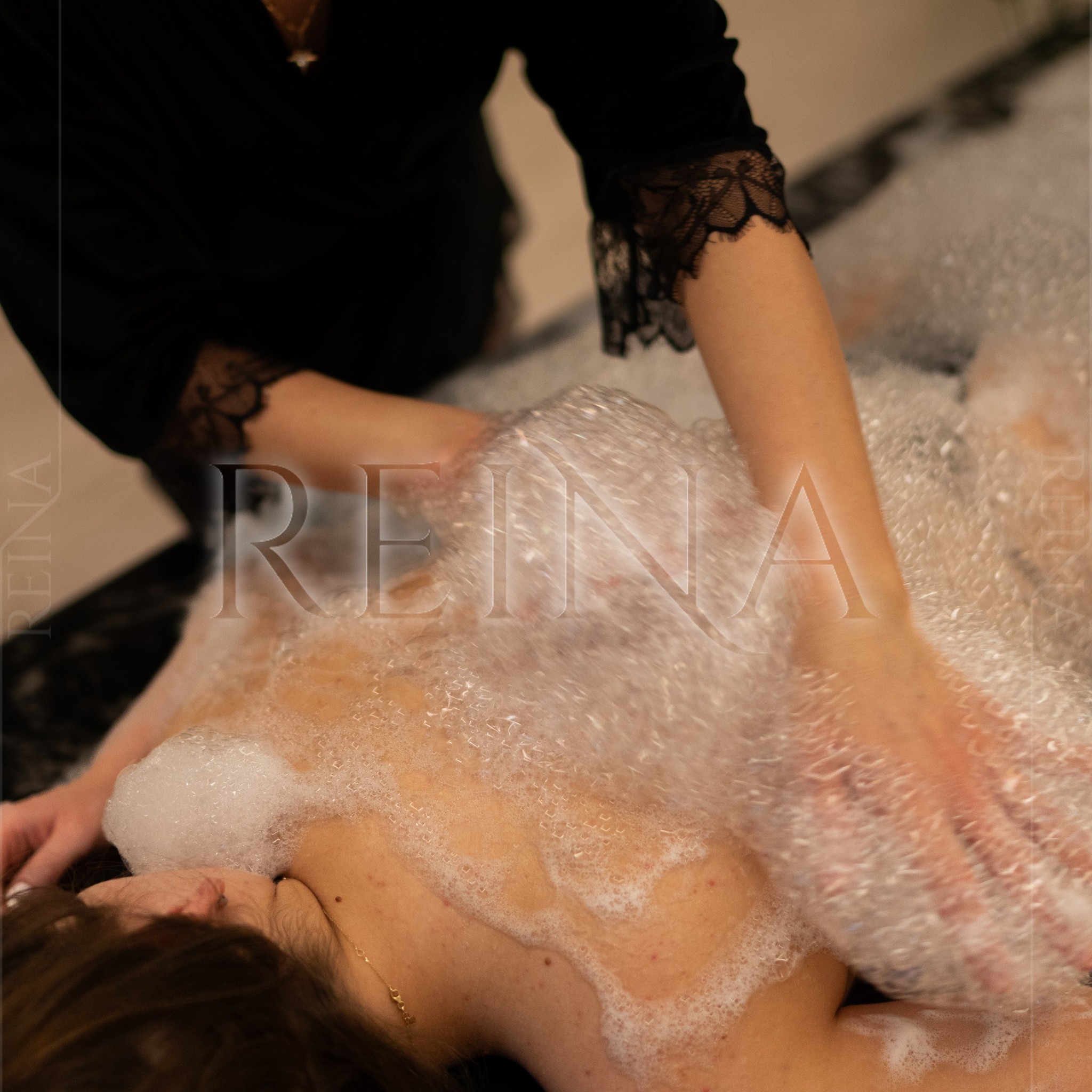 Kobieta relaksuje się podczas zabiegu z pianą, wykonywanego przez masażystkę w czarnym stroju. Delikatne oświetlenie podkreśla atmosferę odprężenia.