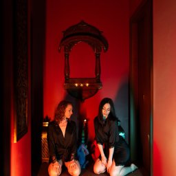 Blissful Massage - Dwie kobiety w szlafrokach klęczą w orientalnym wnętrzu w Warszawie. Klimatyczne oświetlenie świecami i dekoracyjna nisza tworzą atmosferę relaksu.