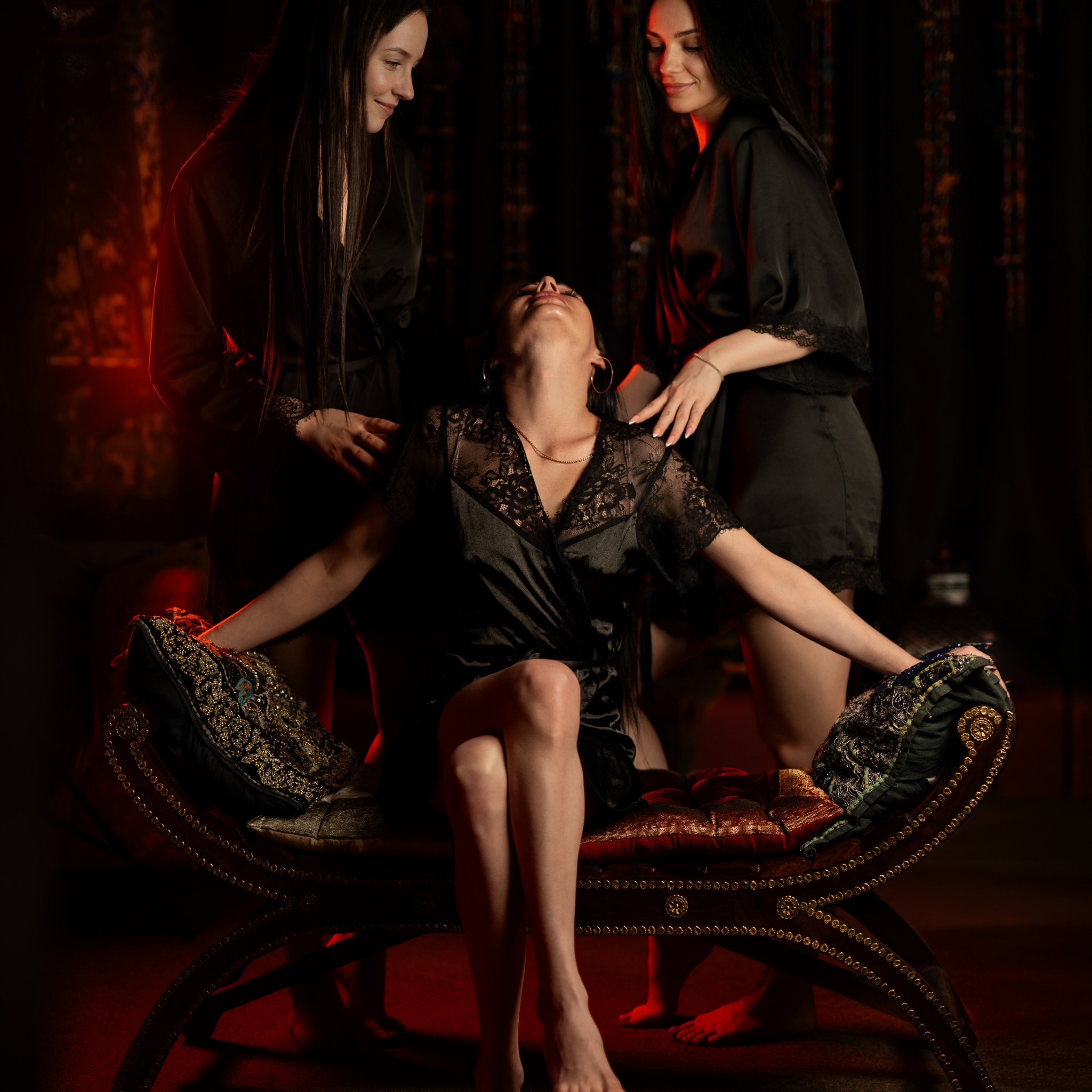 Trzy kobiety w czarnych szlafrokach w ciemnym pomieszczeniu z czerwonym oświetleniem. Dwie stoją i dotykają ramion siedzącej na zdobionej ławce kobiety.