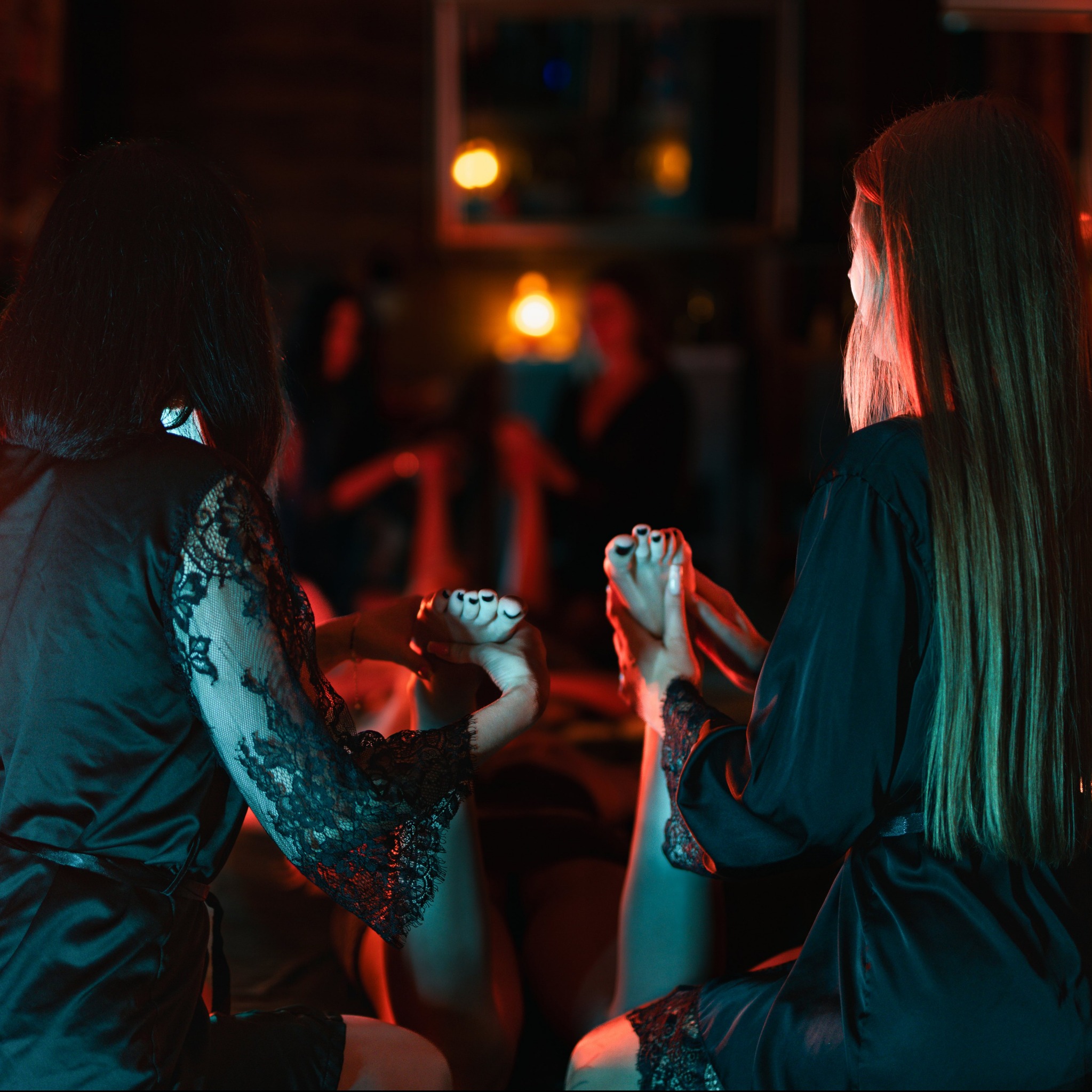 Dwie kobiety w szlafrokach wykonują masaż stóp w klimatycznym, zaciemnionym pomieszczeniu z czerwonym i niebieskim oświetleniem. Widoczne dłonie i stopy klientki.