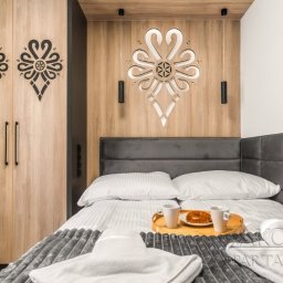 ArchiDESIGN STUDIO PROJEKTOWE arch. Maciej Tubielewicz - Sypialnia z drewnianą zabudową, zdobiona folkowymi wzorami. Nad łóżkiem dekoracja z podświetleniem. Na łóżku taca z kawą i pączkiem. Projekt sypialni w stylu góralskim.