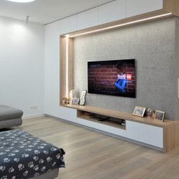 ArchiDESIGN STUDIO PROJEKTOWE arch. Maciej Tubielewicz - Minimalistyczna zabudowa RTV z betonem architektonicznym i LED. Jasne drewno i biel szafek. Telewizor na ścianie. Nowoczesny salon z szarą sofą i drewnianą podłogą.
