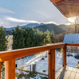 ArchiDESIGN STUDIO PROJEKTOWE arch. Maciej Tubielewicz - Drewniany balkon z przeszkloną balustradą, widok na ośnieżone góry i las. Słoneczny dzień, błękitne niebo z delikatnymi chmurami. Elementy drewniane w ciepłym odcieniu.