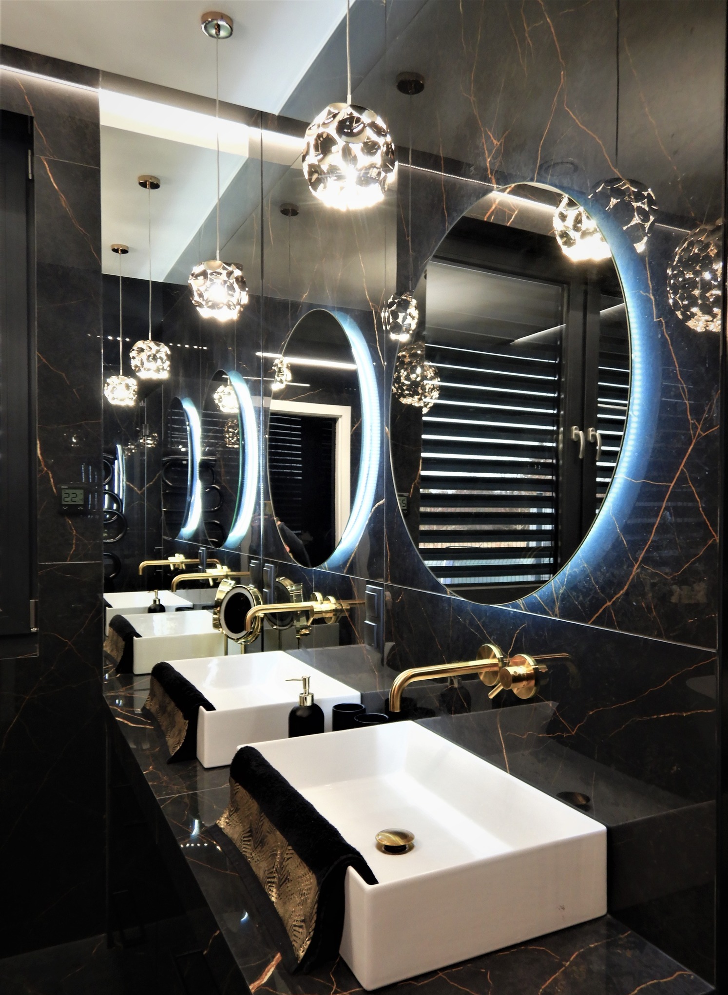 Elegancka łazienka z czarnym marmurem, złotą armaturą i okrągłymi lustrami z podświetleniem LED. Dwie białe umywalki i designerskie lampy wiszące.
