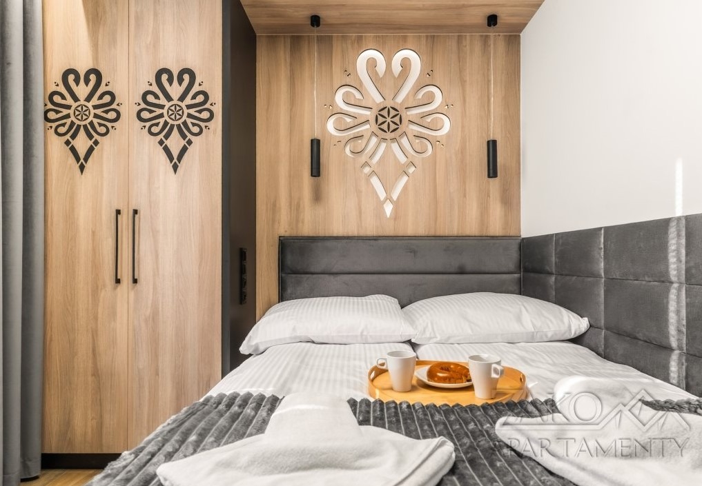 Sypialnia z drewnianą zabudową, zdobiona folkowymi wzorami. Nad łóżkiem dekoracja z podświetleniem. Na łóżku taca z kawą i pączkiem. Projekt sypialni w stylu góralskim.