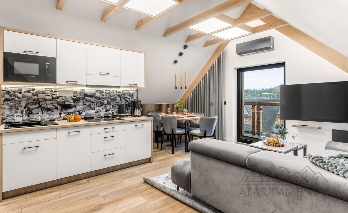 Jasna kuchnia z salonem na poddaszu: białe szafki, drewniane belki stropowe, okno z widokiem na las. Nowoczesny design z unikalnym backsplashiem z motywem zdjęć.