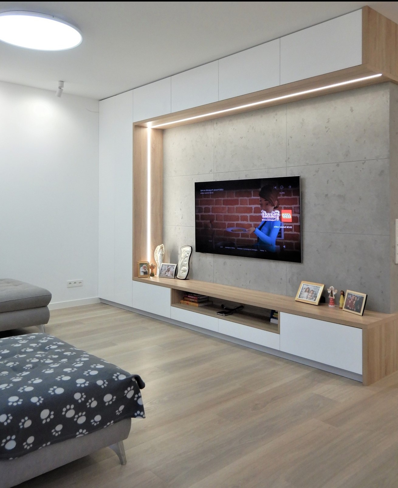 Minimalistyczna zabudowa RTV z betonem architektonicznym i LED. Jasne drewno i biel szafek. Telewizor na ścianie. Nowoczesny salon z szarą sofą i drewnianą podłogą.