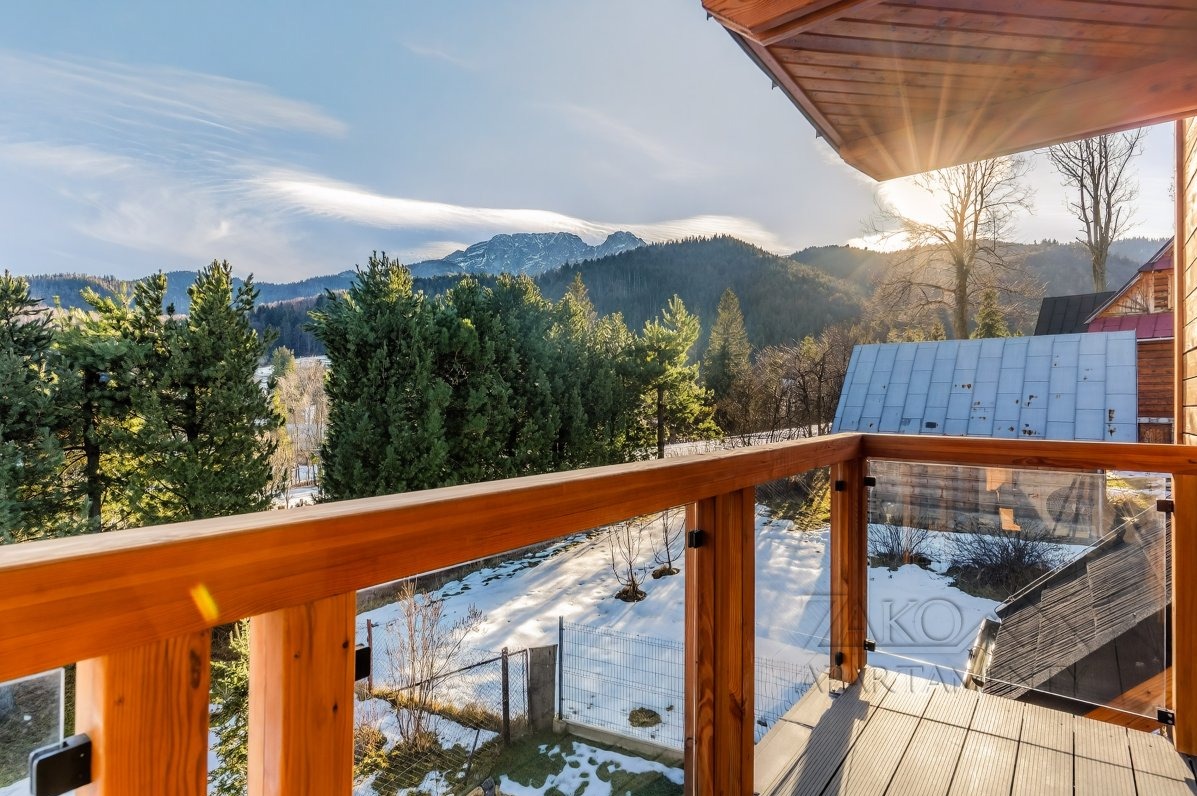 Drewniany balkon z przeszkloną balustradą, widok na ośnieżone góry i las. Słoneczny dzień, błękitne niebo z delikatnymi chmurami. Elementy drewniane w ciepłym odcieniu.
