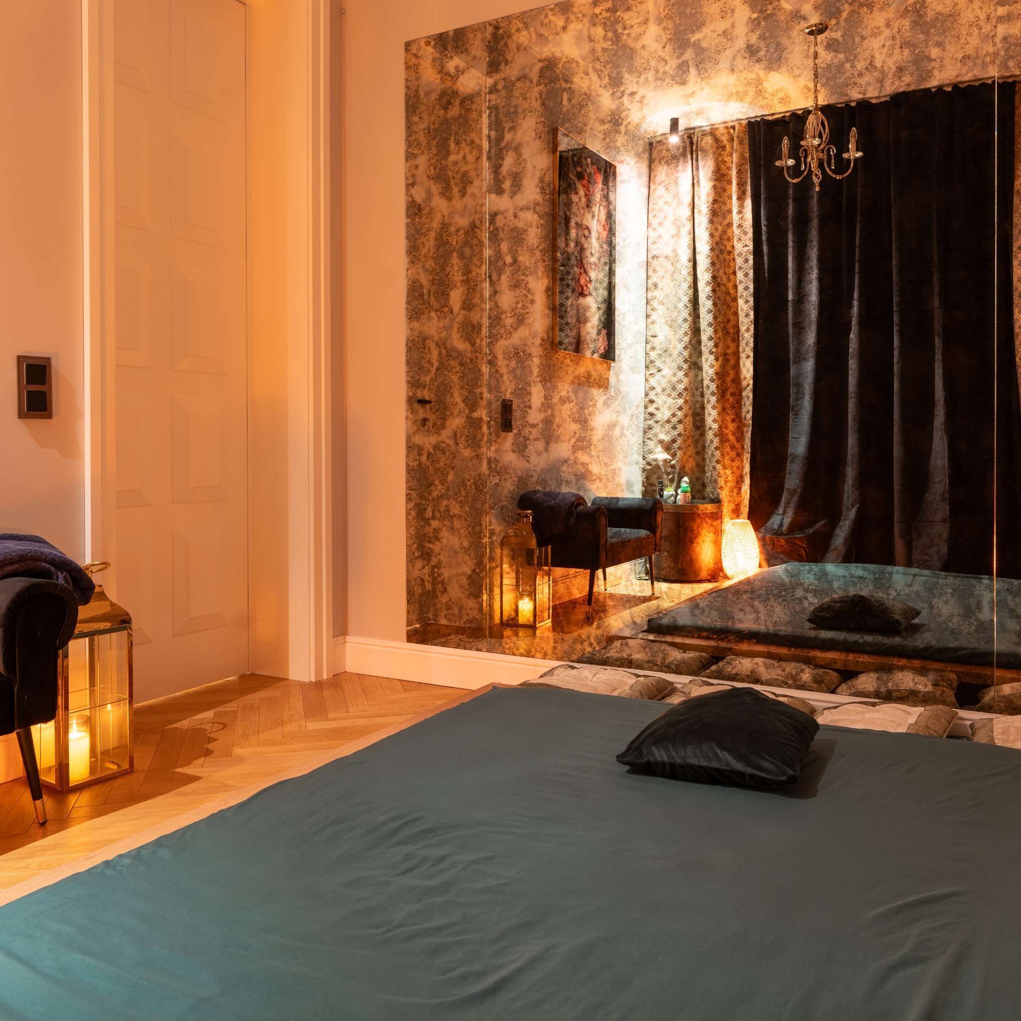 Luksusowe wnętrze gabinetu z łóżkiem do zabiegów, lustrzaną ścianą i dekoracyjnym oświetleniem. Widoczny fotel, lampion ze świecą i zasłony. Ciepłe, relaksujące światło.