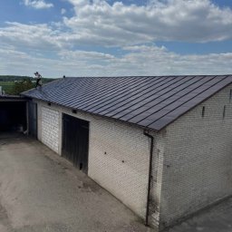 Usługi Dachowe - Ciemny dach z blachy na jasnym budynku z cegły. Na dachu stoi osoba. Widoczne rynny i błękitne niebo z chmurami. Ujęcie z góry.