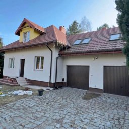 Usługi Dachowe - Odnowiona elewacja domu z brązowymi bramami garażowymi i kostką brukową. Widoczne narzędzia malarskie sugerują świeżo zakończone prace.