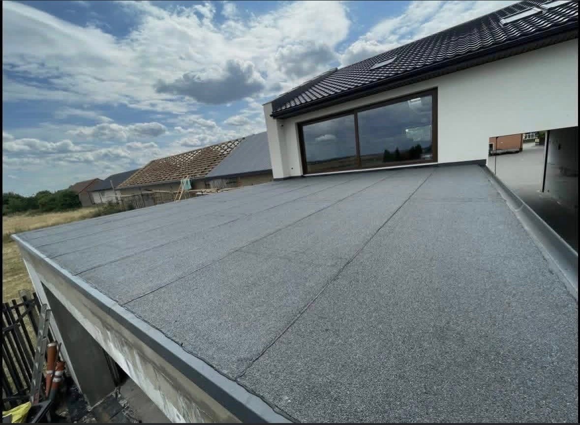 Szczelny, płaski dach pokryty papą termozgrzewalną na nowoczesnym domu z dużym oknem. Widoczne detale wykończenia i sąsiednie budynki w tle.