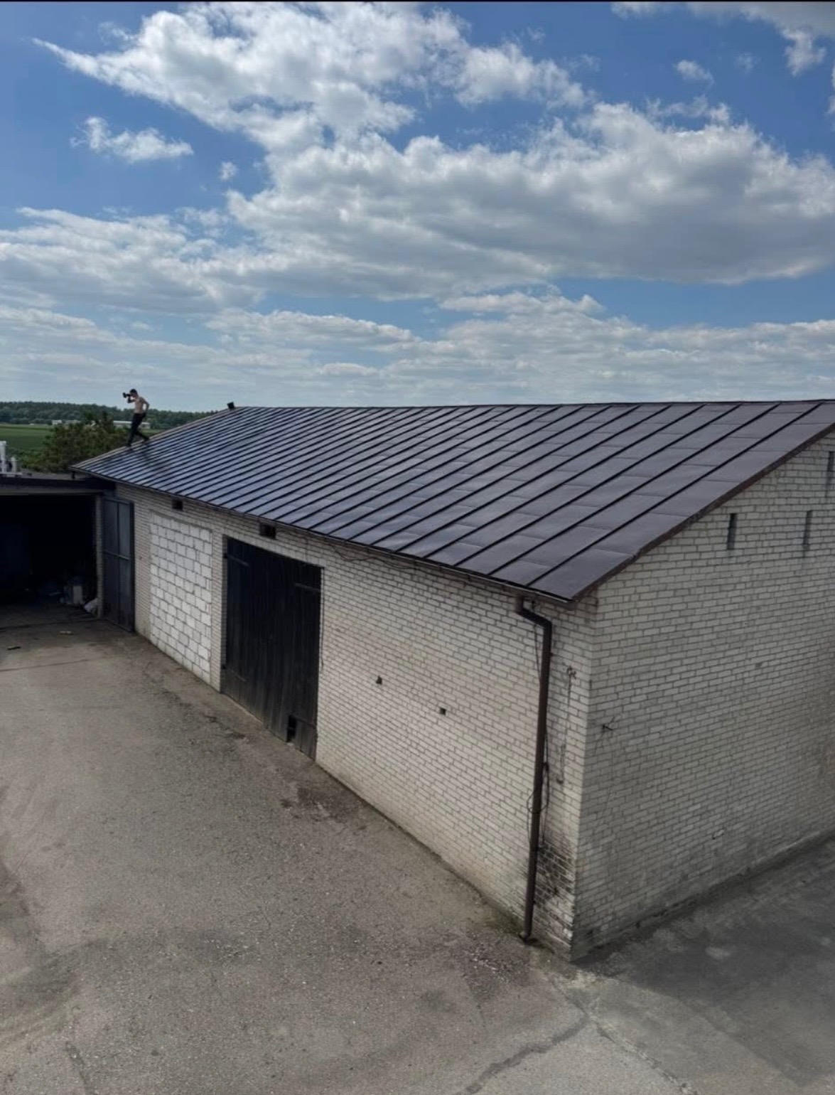 Ciemny dach z blachy na jasnym budynku z cegły. Na dachu stoi osoba. Widoczne rynny i błękitne niebo z chmurami. Ujęcie z góry.