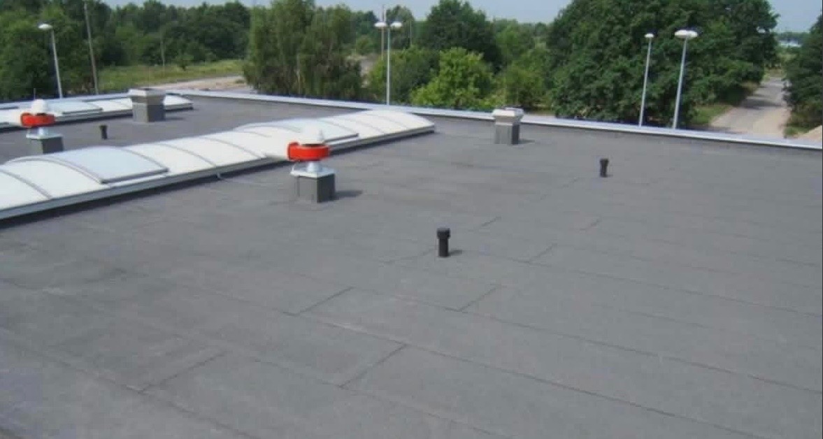 Płaski dach pokryty papą z elementami wentylacyjnymi i świetlikami. Widok z góry, w tle drzewa i latarnie.