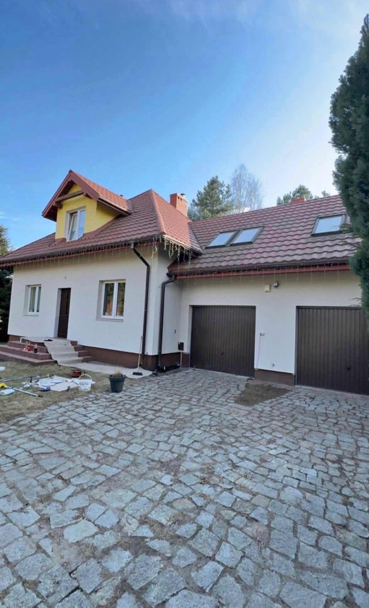 Odnowiona elewacja domu z brązowymi bramami garażowymi i kostką brukową. Widoczne narzędzia malarskie sugerują świeżo zakończone prace.