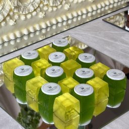 LUO SPÓŁKA Z OGRANICZONĄ ODPOWIEDZIALNOŚCIĄ - Żółto-zielone galaretki w kształcie kostek, z logo firmy cateringowej, ułożone na lustrzanej tacy. W tle kremowy tort z dekoracjami.