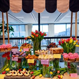 LUO SPÓŁKA Z OGRANICZONĄ ODPOWIEDZIALNOŚCIĄ - Stół z deserami pod markizą w biurze: babeczki, ciasta, cake pops i tulipany w wazonach. Słodki bufet na tle okien biurowca.