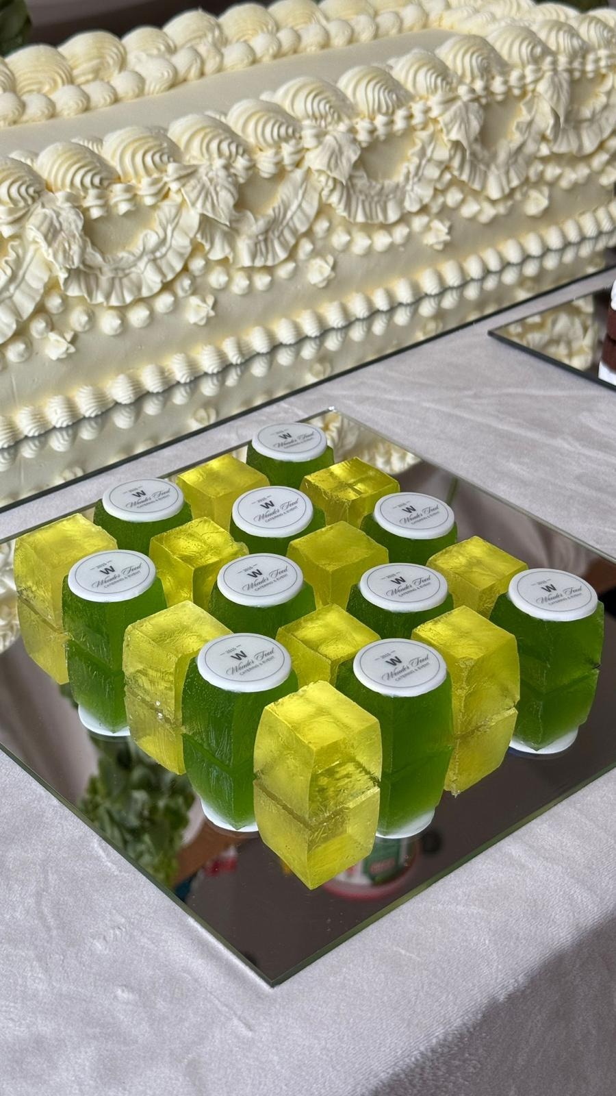 Żółto-zielone galaretki w kształcie kostek, z logo firmy cateringowej, ułożone na lustrzanej tacy. W tle kremowy tort z dekoracjami.