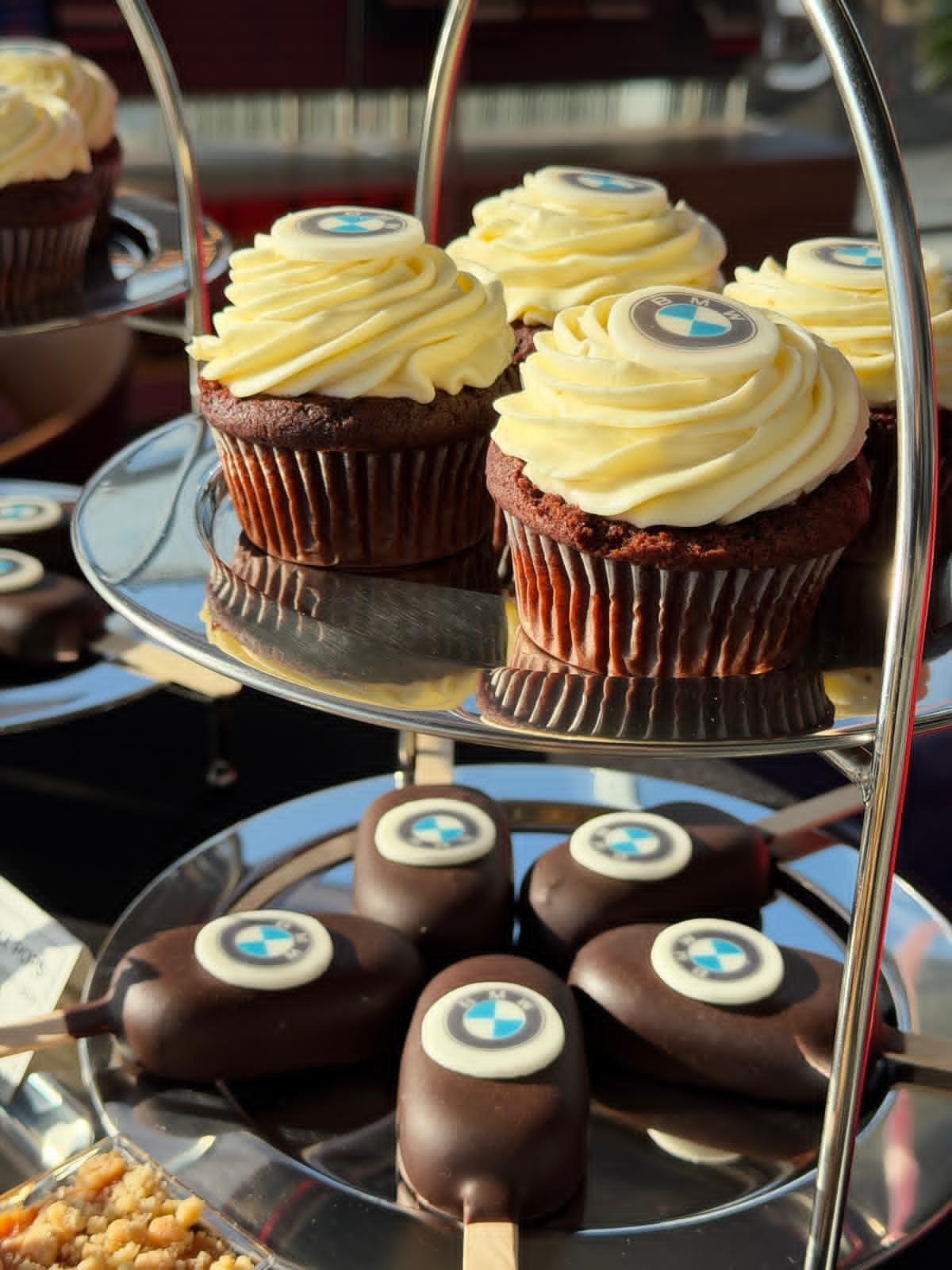 Elegancki, srebrny etażer z czekoladowymi babeczkami z kremem i lodowymi ciastkami na patyku, ozdobionymi logo. Słodki poczęstunek na firmowe wydarzenie.