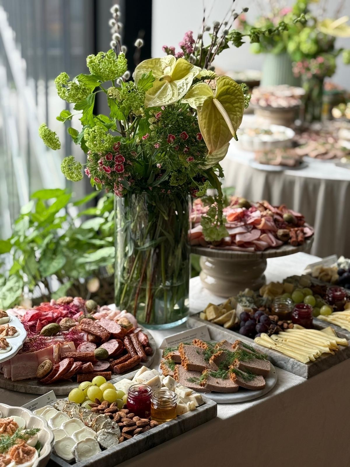 Bogato zastawiony stół z przekąskami: sery, wędliny, owoce, pasztet i kwiatowe dekoracje w szklanym wazonie. Elegancki catering na przyjęcie.