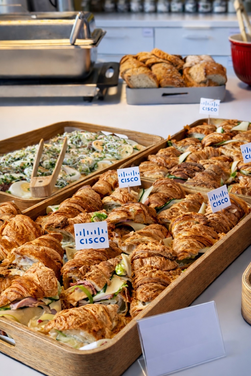 Kruche croissanty z nadzieniem i sałatka w drewnianych korytkach, oznaczone firmowymi etykietami Cisco, na białym stole. W tle pieczywo i naczynia.