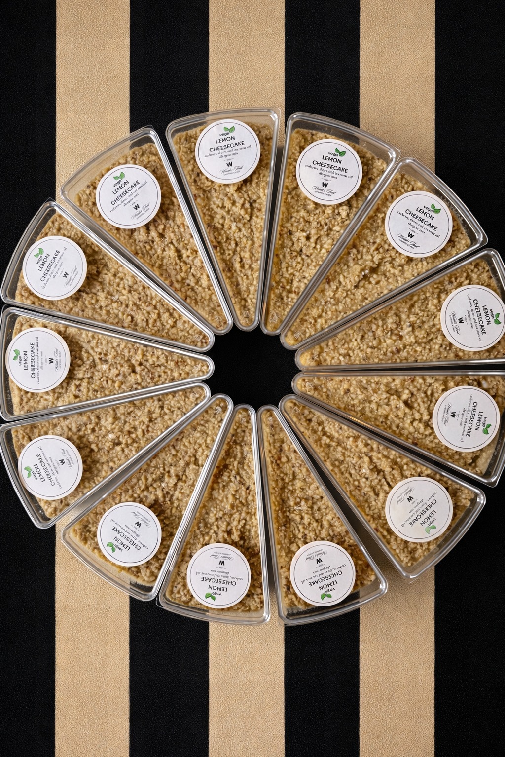 Okrąg z porcji wegańskiego sernika cytrynowego w trójkątnych opakowaniach, z etykietami. Czarno-złote tło w paski. Widok z góry, ostre światło.