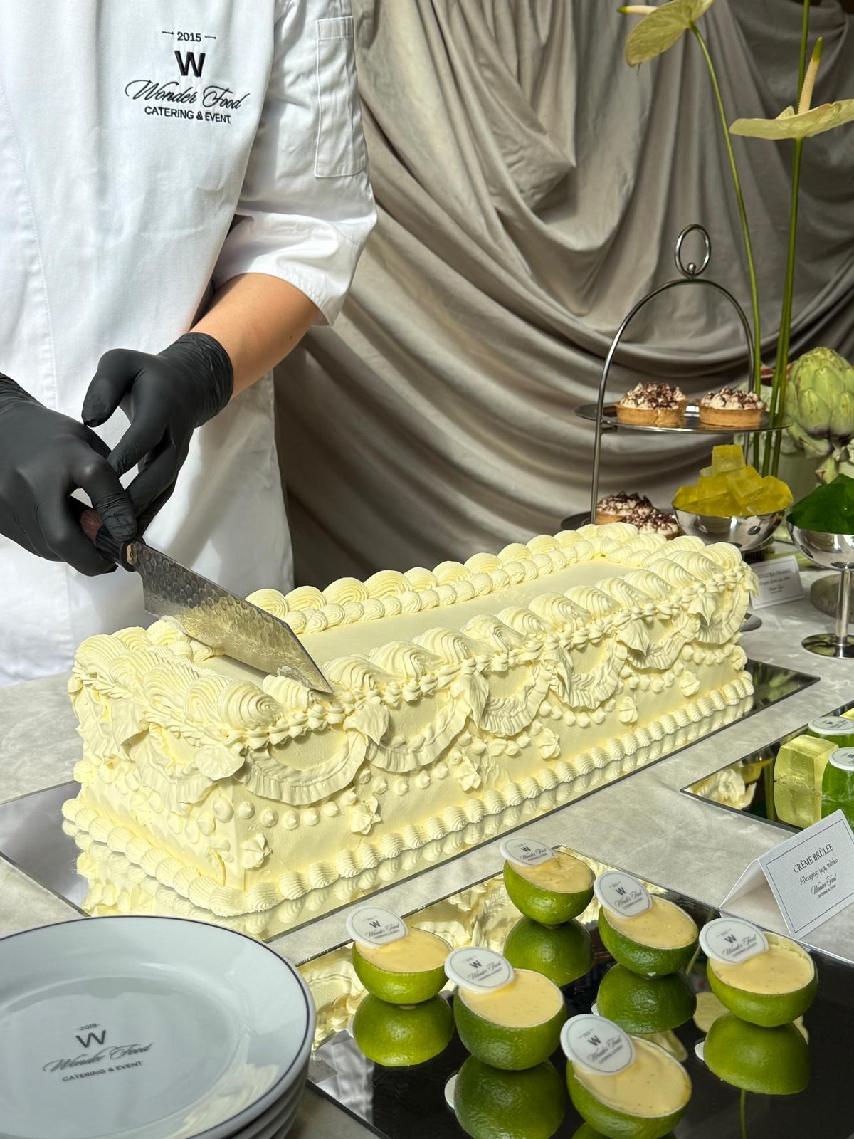 Kucharz w czarnych rękawiczkach kroi ozdobny tort na lustrzanej tacy. W tle desery, kwiaty i drapowana tkanina. Elegancki catering na imprezę.