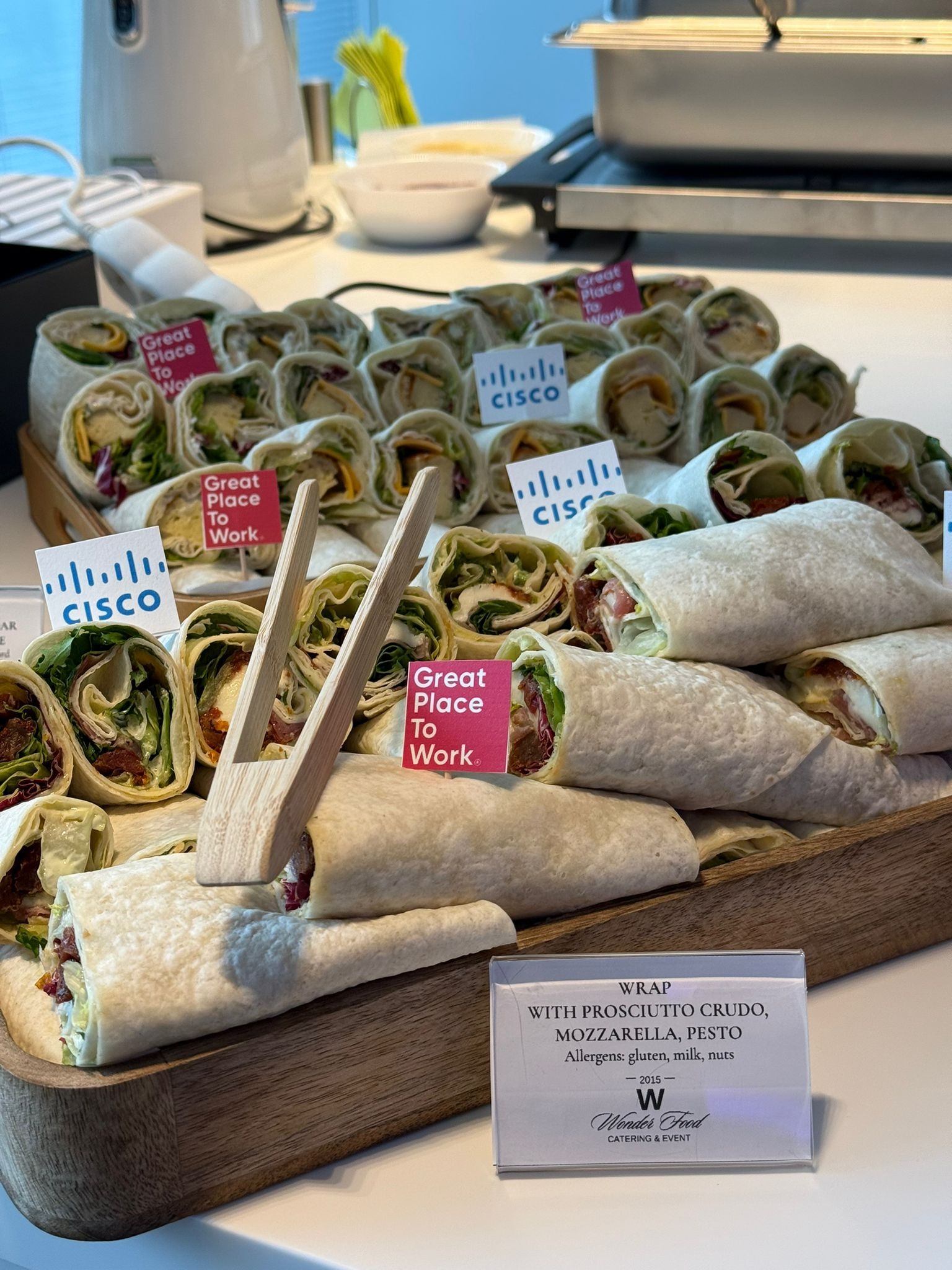 Wrapy z prosciutto crudo, mozzarellą i pesto na drewnianej tacy, z etykietami 'Cisco' i 'Great Place to Work', oferowane przez Wonder Food Catering & Event.