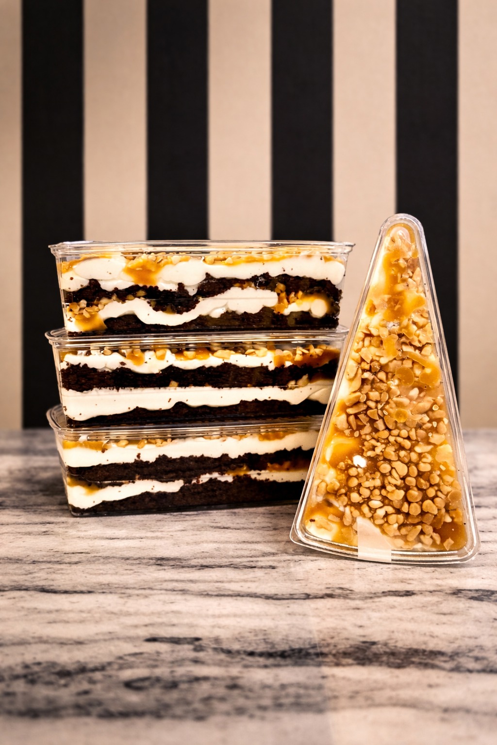 Trzy prostokątne desery warstwowe i deser w kształcie choinki, w przezroczystych pojemnikach, na marmurowym blacie, z czarno-beżowym tłem w paski.