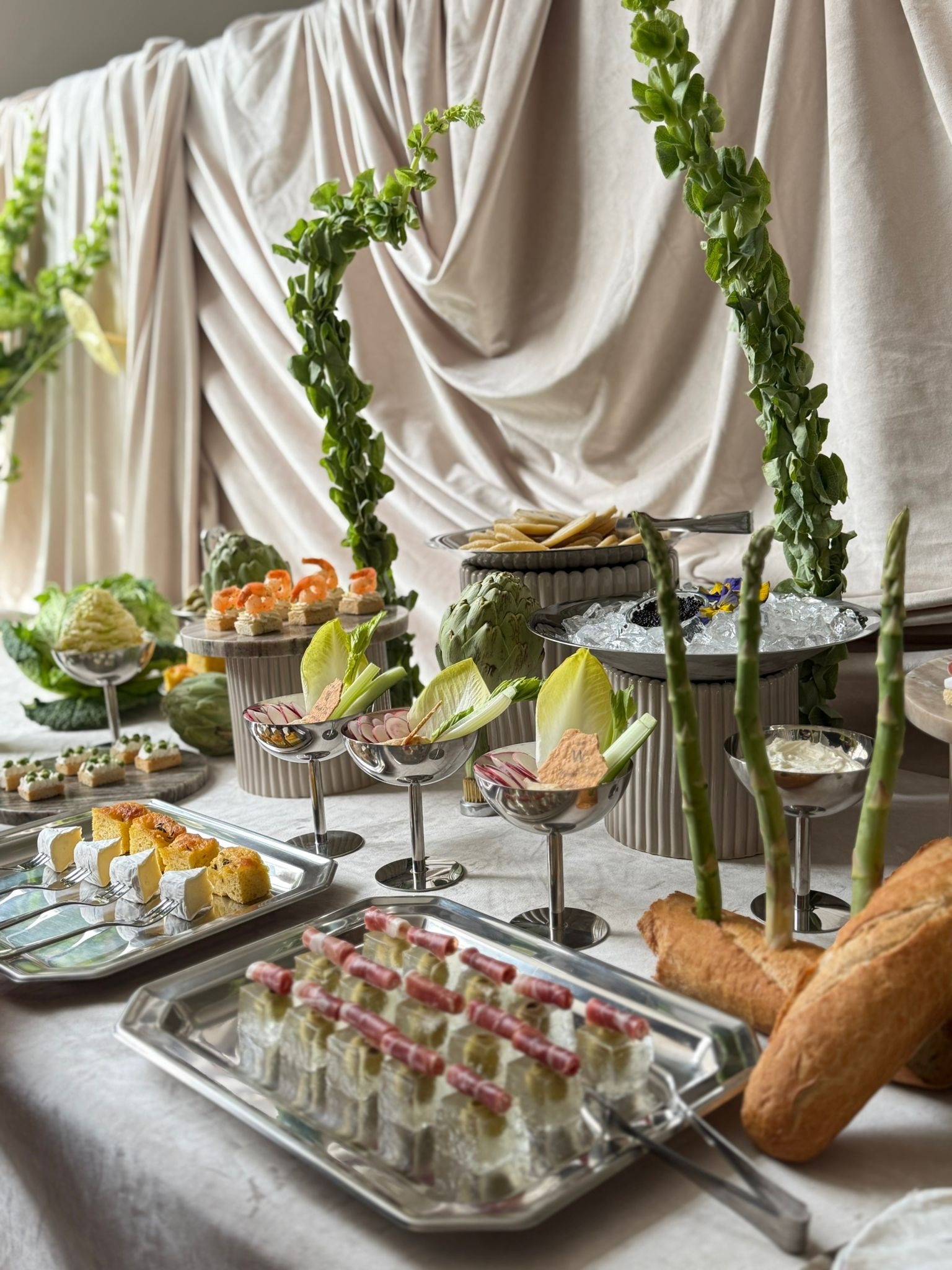 Elegancki stół cateringowy z wykwintnymi przekąskami, dekoracjami roślinnymi i srebrnymi naczyniami na tle drapowanej tkaniny. Widoczne szparagi, sery i wędliny.