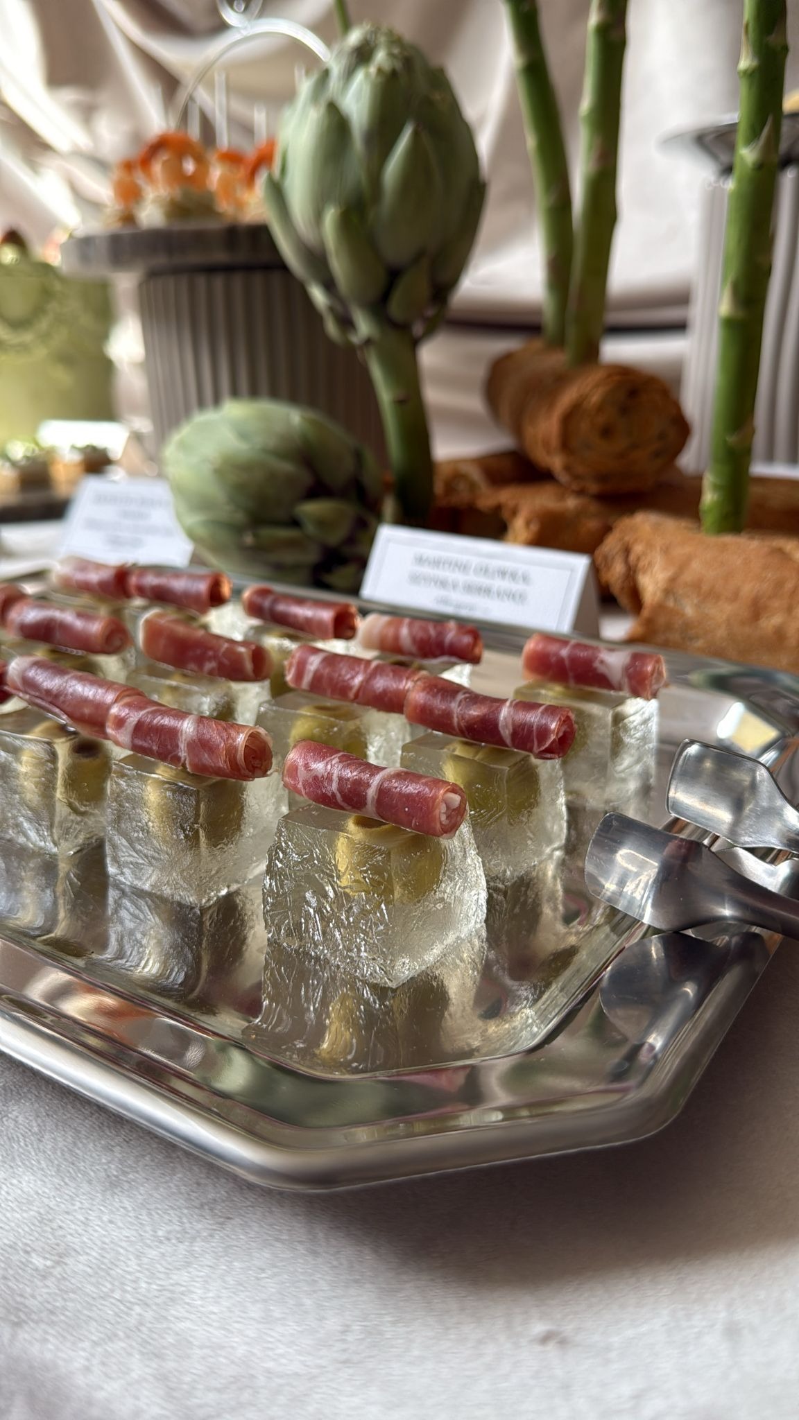Elegancki catering: kostki lodu z oliwkami i szynką na srebrnej tacy, w tle artystycznie ułożone przekąski i karczochy. Wyrafinowane detale i dbałość o prezentację.