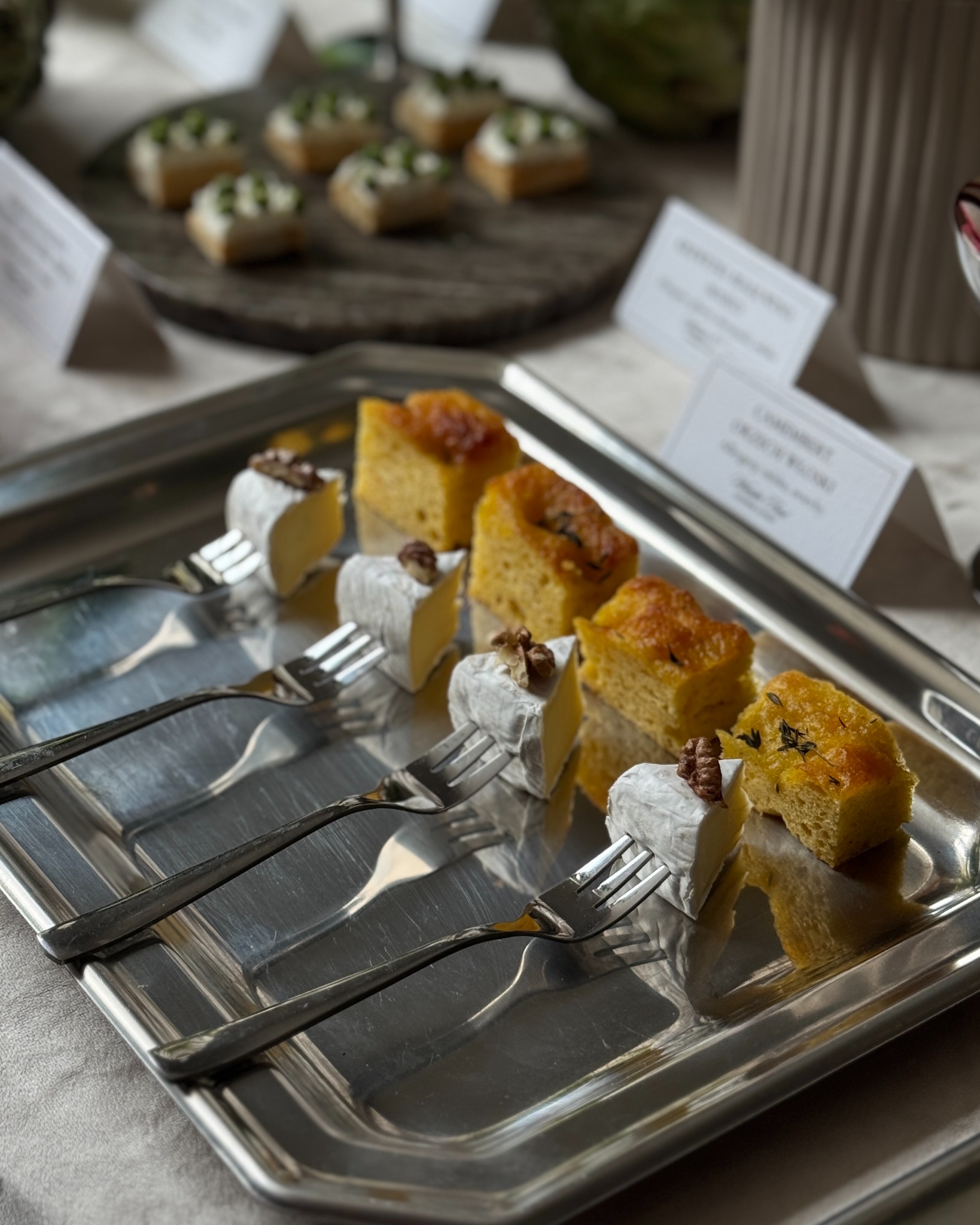 Srebrna taca z serami brie z orzechami i kawałkami ciasta na widelcach, elegancko podane, w tle inne przekąski i etykiety z nazwami.