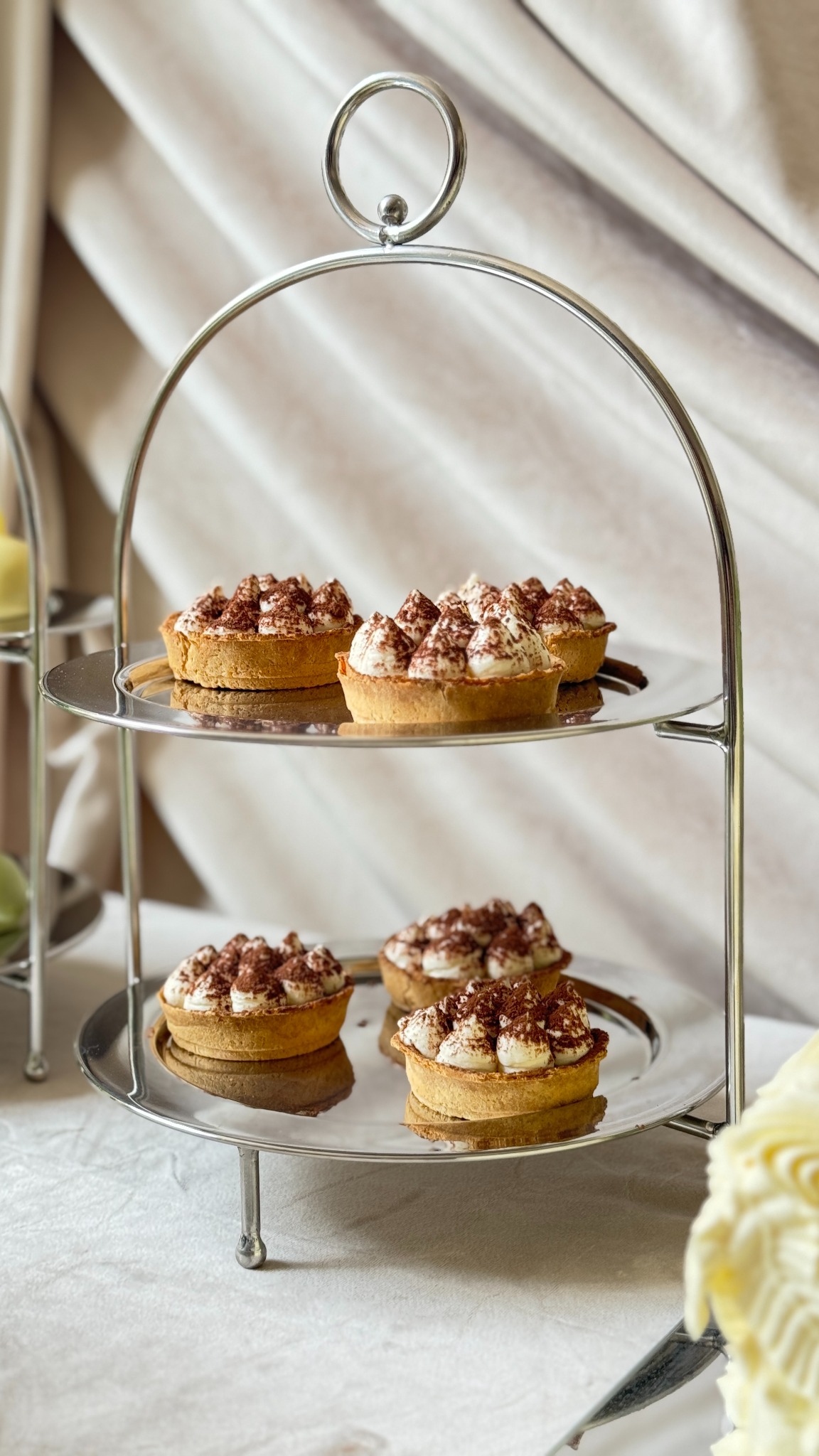 Dwupoziomowy srebrny etażer z mini tartaletkami tiramisu na białym obrusie i kremowym tle. Elegancki deser na przyjęcie.