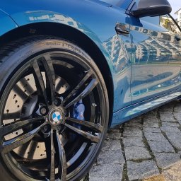 Izdebski Studio - Auto Detailing Warszawa - Błyszczące, czarne felgi BMW z niebieskimi zaciskami hamulcowymi, po detailingu. Widoczny fragment karoserii w odcieniu turkusowym, odbijający budynki.