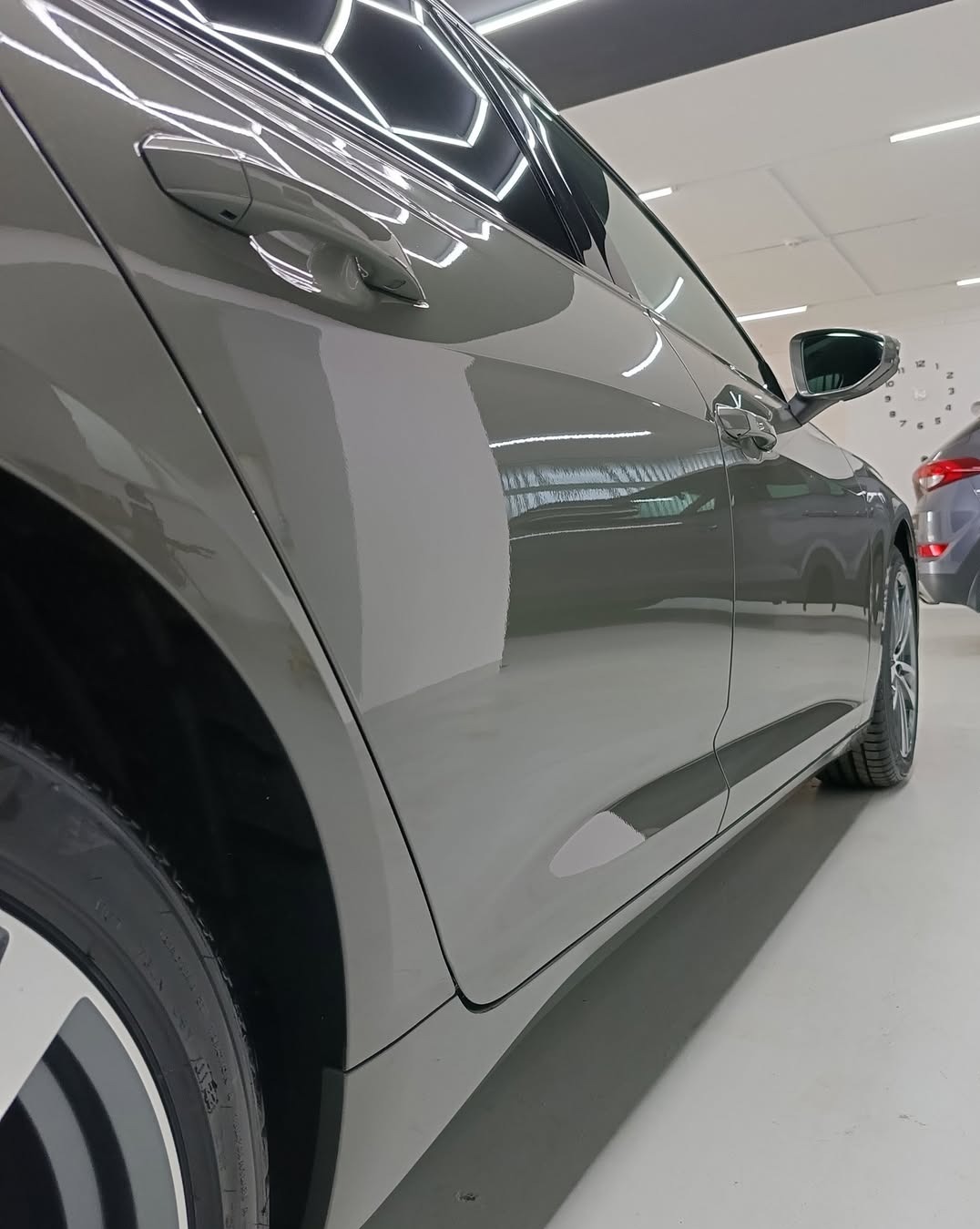Błyszczący, szary samochód po detailingu, z efektem lustrzanego odbicia na lakierze, widoczny fragment opony i felgi, oświetlenie LED w tle.
