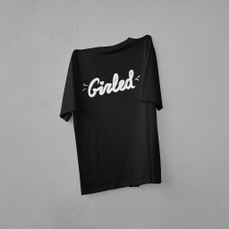 Dawid Zapart - Czarna koszulka z białym napisem 'Girled' zawieszona na szarej ścianie. Minimalistyczny design, stonowana kolorystyka, proste tło. Prezentacja nadruku na odzieży.