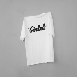 Dawid Zapart - Biała koszulka z czarnym napisem 'Girled' zawieszona na szarej ścianie. Minimalistyczny design, proste tło, ostre światło podkreśla teksturę materiału.