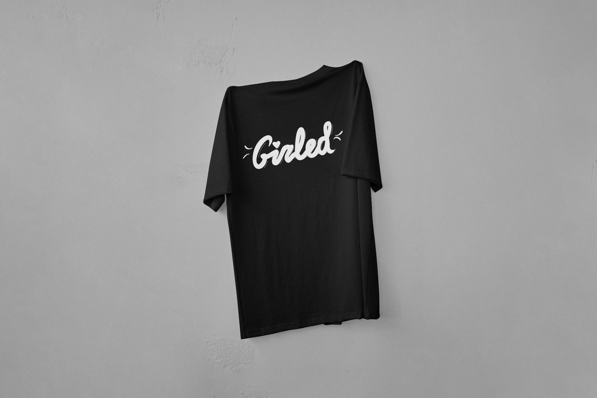 Czarna koszulka z białym napisem 'Girled' zawieszona na szarej ścianie. Minimalistyczny design, stonowana kolorystyka, proste tło. Prezentacja nadruku na odzieży.