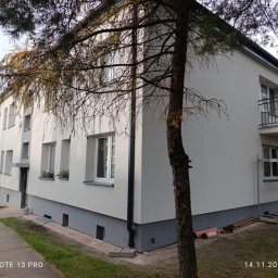 ALFA BUD MYKHAILO SAMOILICH - Odnowiona elewacja budynku wielorodzinnego z szarą podmurówką i białymi ścianami, widoczna z boku, z drzewem na pierwszym planie i rusztowaniem przy balkonie.