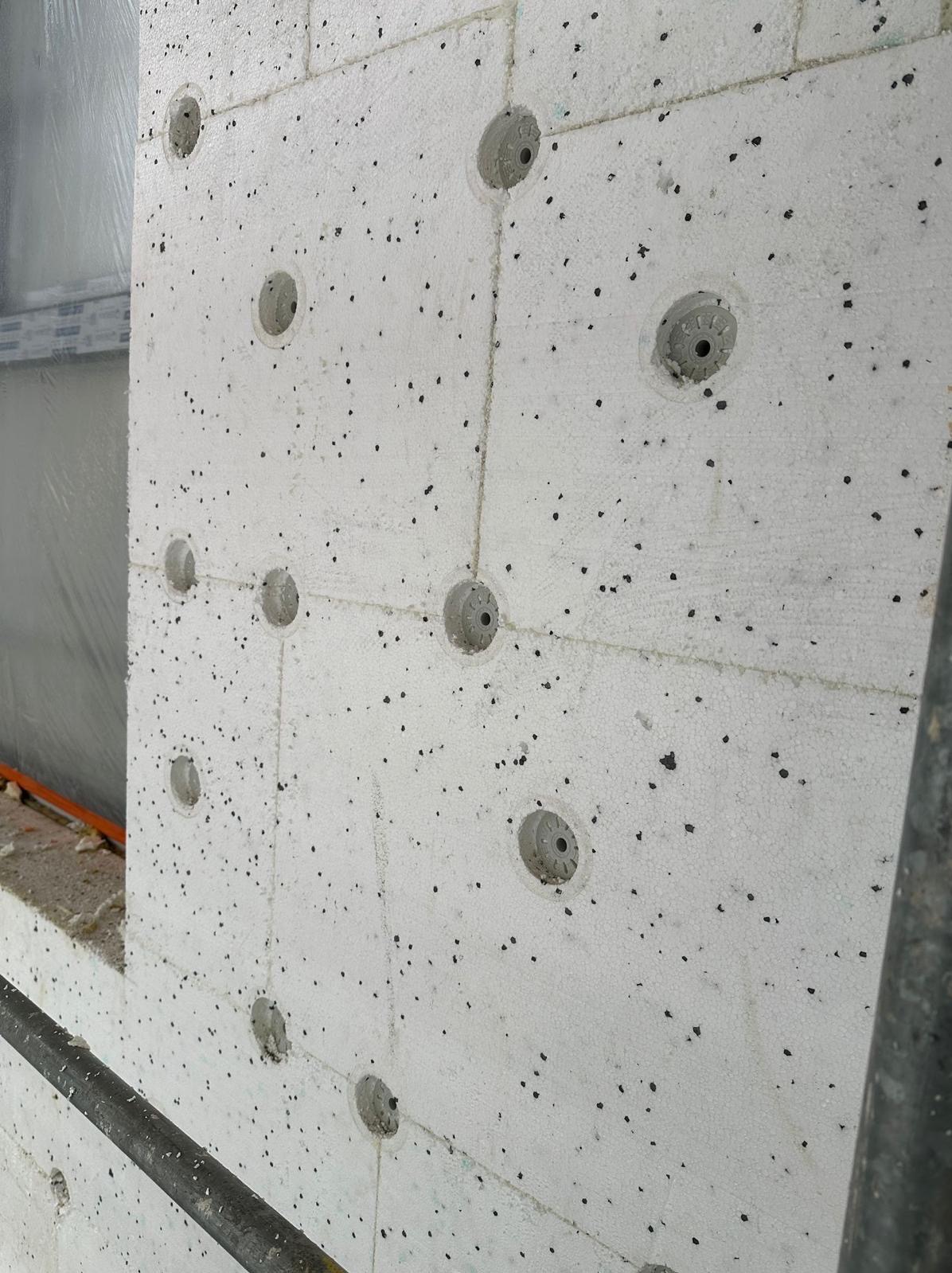 Ocieplona ściana budynku z widocznymi otworami na kołki mocujące, pokryta styropianem z grafitowym wzorem. Fragment okna w tle. Widoczny fragment rusztowania.